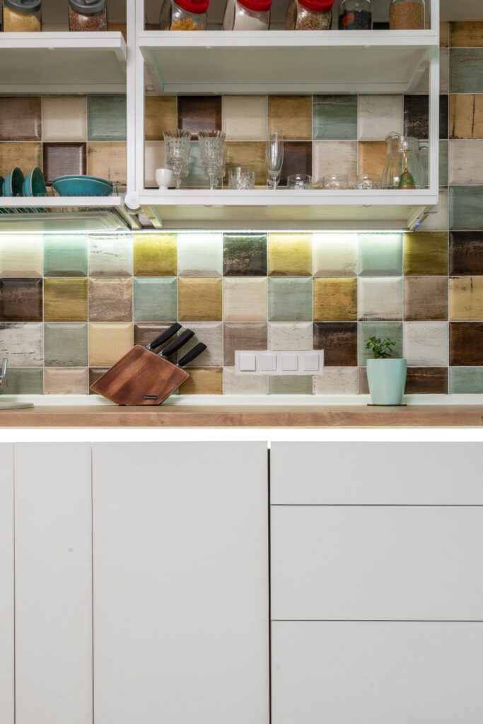 Interior of modern light kitchen furnished with wooden counter and white cupboards and shelves decorated with various dishware and cups with tiled wall